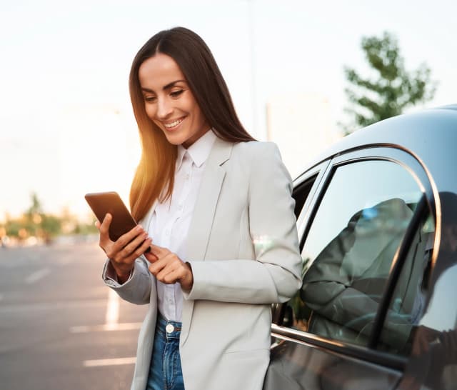 Bild einer gut gekleideten Frau mit Handy in der Hand, neben einem Auto.